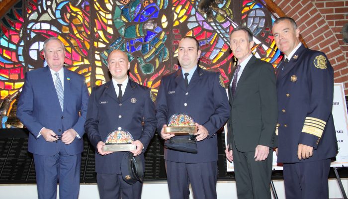 two paramedics hold awards while standing in front of stained glass window with three men in suits
