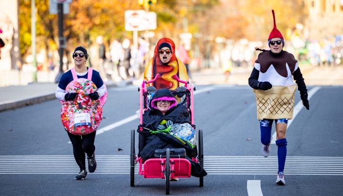 Three participants in the Philadelphia Marathon are dressed up in costume. One is a donut, one is a hotdog, and one is an ice cream cone. The participant in the hot dog costume is pushing a wheel chair participant.