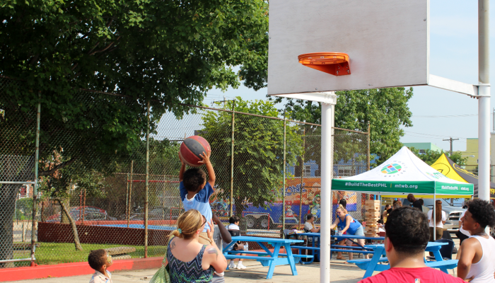 A woman lifts up a young child, so he shoot the basketball into the hoop.