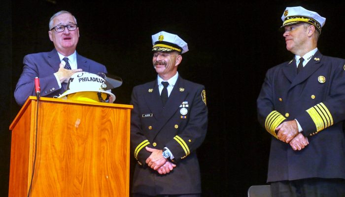 mayor stands at podium holding firefighter helmet as two fire chiefs in dress uniforms look at him