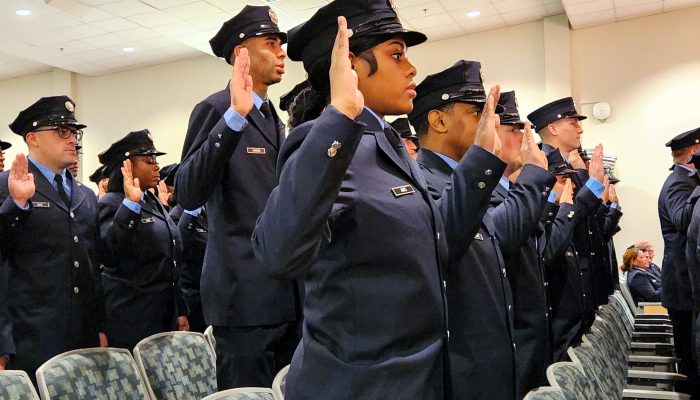 viewed from the side, firefighters in dress uniforms stand in auditorium with their right hand raised