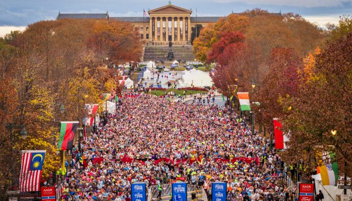 The morning of the Philadelphia Marathon. Tens of thousands of runners are lined up at the start. The tree foliage is orange and red. The grand steps of the Art Museum are in the backgound.