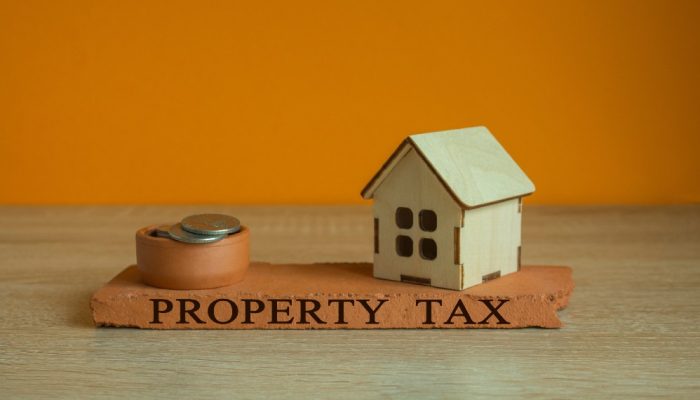 A wooden house and stack of coins on a wooden plank with Property Tax written on it