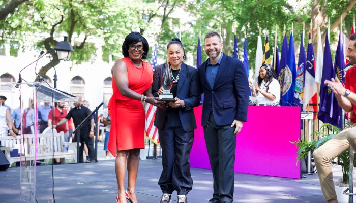 Mayor Cherelle Parker stands on stand beside a podium with flags lining the backdrop as she poses with native Philadelphian Dawn Staley as Staley holds the One Philly Award presented to her by the mayor. Next to them also posing in the photo is representative from Wawa Welcome America and audience members can be seen in the background as well. 