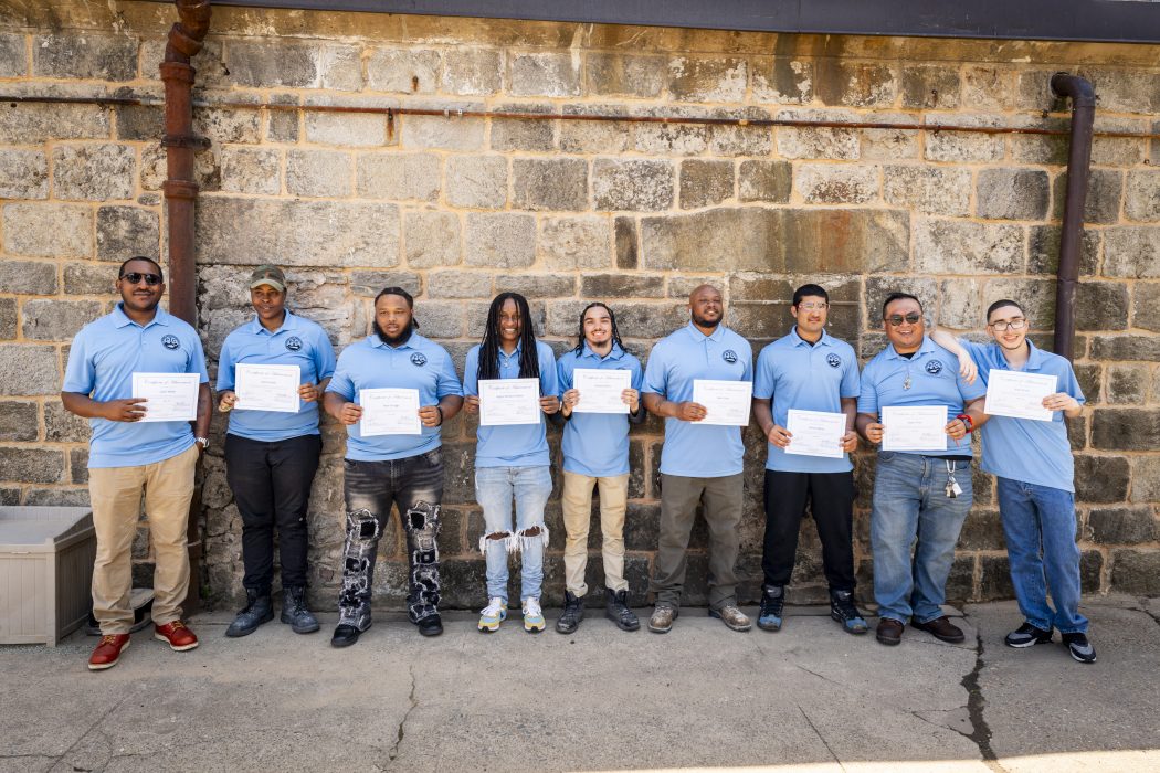 Ten masonry academy graduates in blue shirts holding diplomas in front of a stone wall.