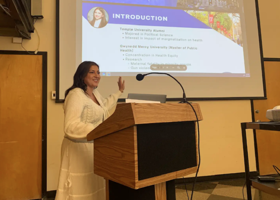 A VISTA member presents at a podium, with a projected slide about her education and research interests in public health and health equity behind her.