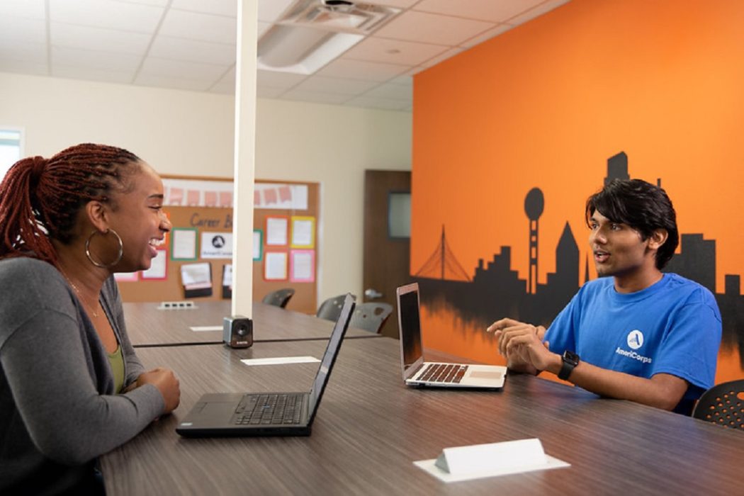 Two individuals seated at a table across from one another, each using a laptop, engaged in a collaborative work session.