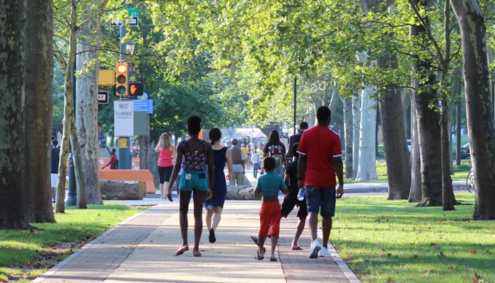 A family walking on the sidewalk on the Benjamin Franklin Parkway in Philadelphia, surrounded by green trees.