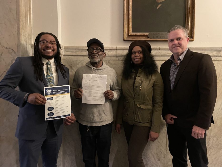 A councilmember holding a Title Clearance Unit flyer and a man holding his deed standing next to the Register of Wills Staff.