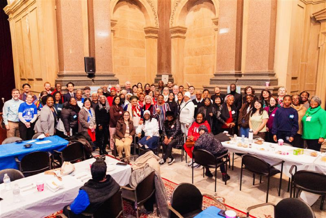 A large group of Community School volunteers pose for a photo in a spacious hall. The setting is lively and warm, with tables set for a communal event.