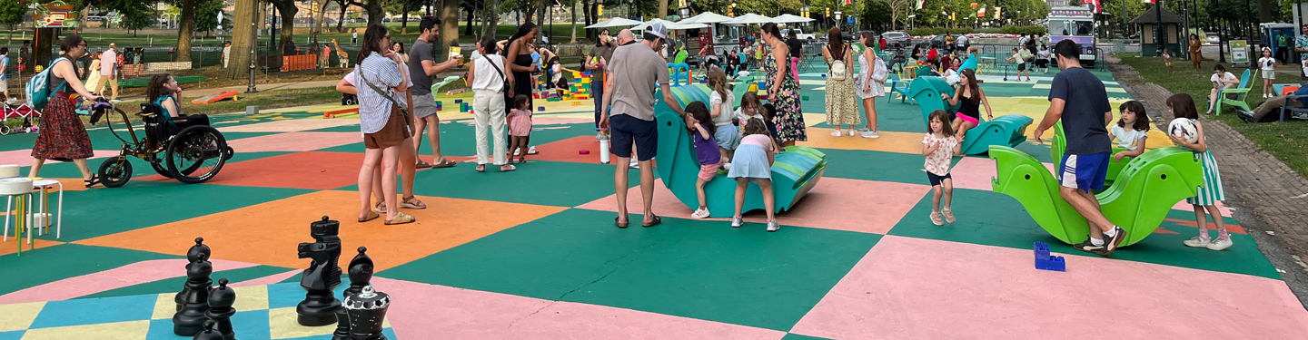 Young kids playing in an open space surrounded by trees with colorful art, giant blocks and chess pieces, and teeter totters.
