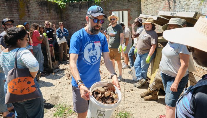 A crowd of people gather around a bucket of compost at a Farm Philly demonstration.