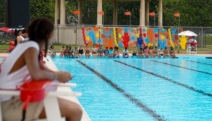 A lifeguard watches a line of young people sit on the edge of a pool.