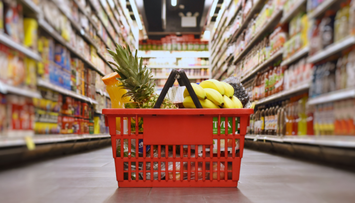 A red shopping basket filled with groceries sits on the floor of a grocery store aisle with full grocery shelves visible on either side.