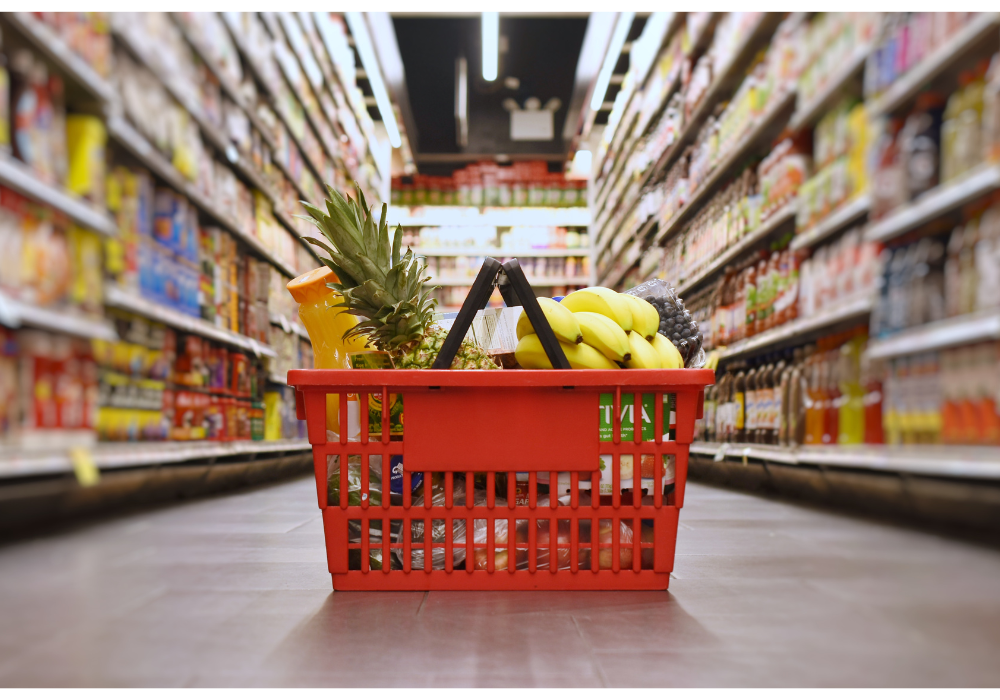 A red shopping basket filled with groceries sits on the floor of a grocery store aisle with full grocery shelves visible on either side.