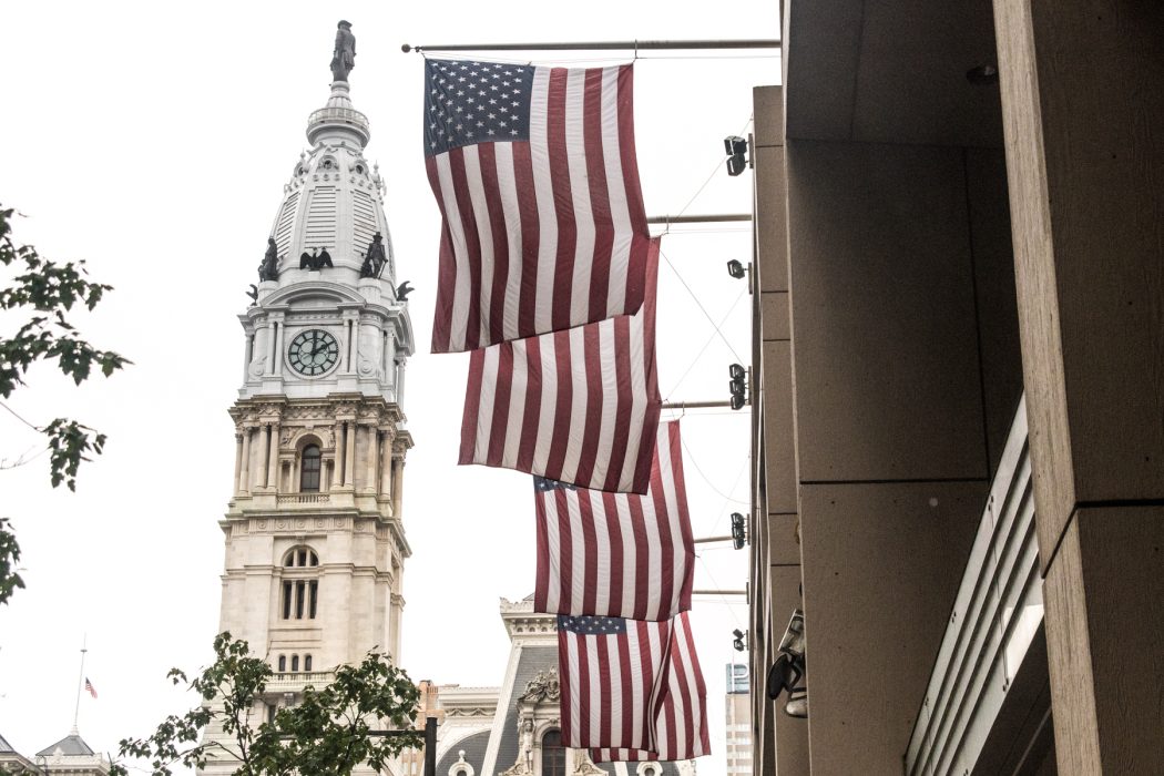 Four American flags hang vertically from the side of a building, with Philadelphia's historic City Hall and its clock tower prominently visible in the background. The statue of William Penn stands atop the tower, and some tree branches are visible in the foreground.