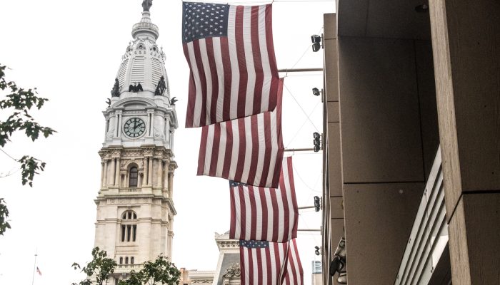 Four American flags hang vertically from the side of a building, with Philadelphia's historic City Hall and its clock tower prominently visible in the background. The statue of William Penn stands atop the tower, and some tree branches are visible in the foreground.