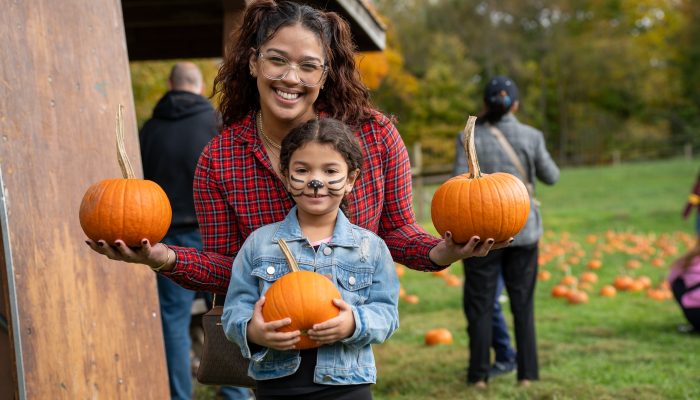 An adult and young child smile and hold up pumpkins in a open field.