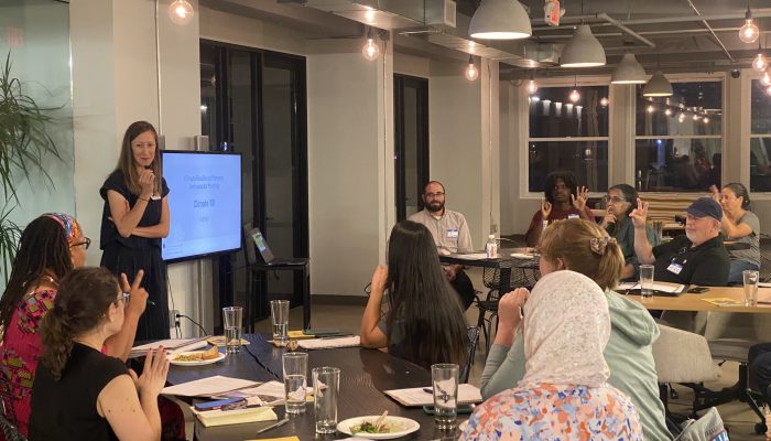 People sitting around round tables and listening as a person standing by a presentation screen speaks