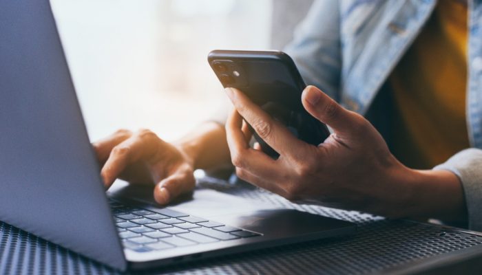 One hand holding a phone, the other browsing a laptop