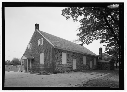 Historical, black and white photo showing an exterior view of the Frankford Preparative Friends Meeting House. This angle shows that one exterior wall is made of brick and another is constructed of stone. In the background is the burial ground.