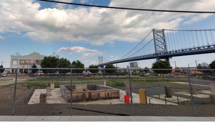A fenced in lot with a foundation under construction. The Benjamin Franklin bridge and a pier building can be seen in the background.