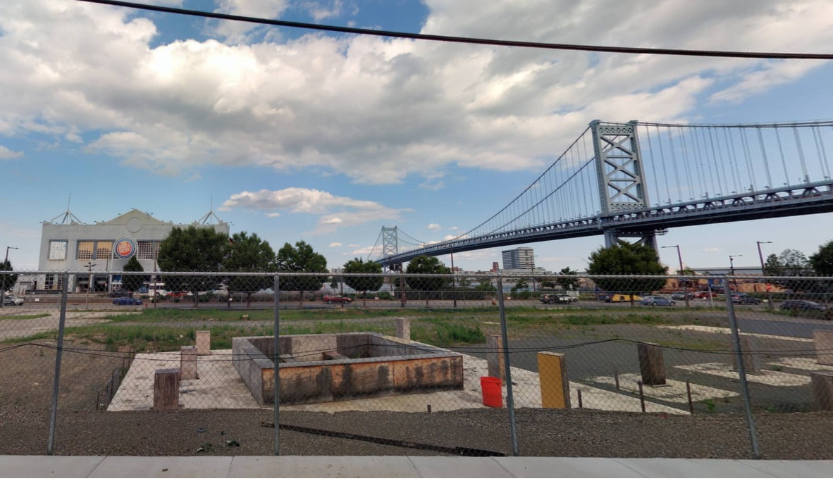 A fenced in lot with a foundation under construction. The Benjamin Franklin bridge and a pier building can be seen in the background.