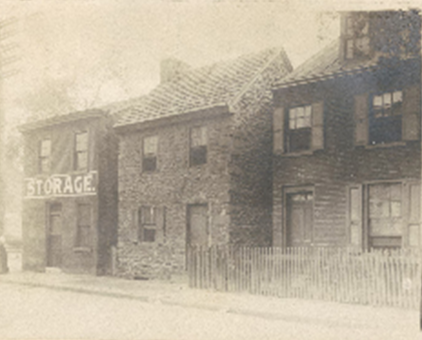 A faded photograph shows a stone house in the center. A warehouse marked “storage” appears at the left of the photo and a house with wood siding appears on the left.