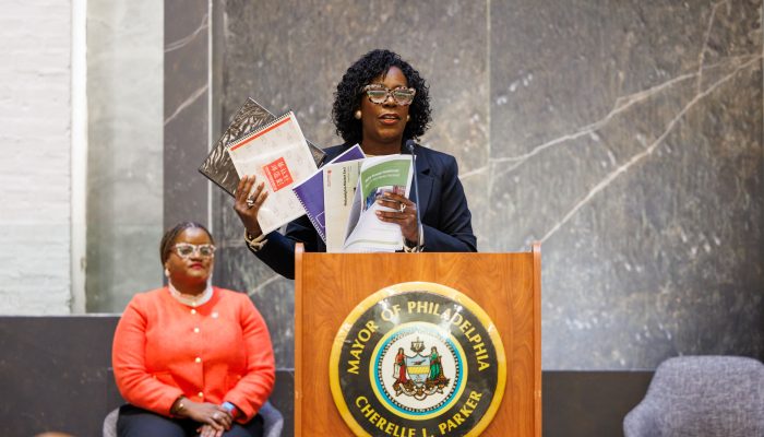 A photo of Mayor Parker speaking at the podium during the November 7, 2025, announcement of the formation of the Market East Advisory Group. She is holding multiple printed and bound copies of old plans for Market East. Sitting behind her is Chief of Staff Tiffany Thurman.