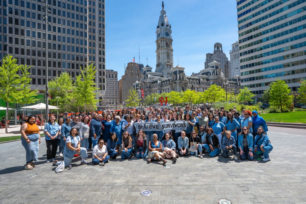 A smiling group wearing denim poses with a banner that says "Believe Survivors" in Love Park