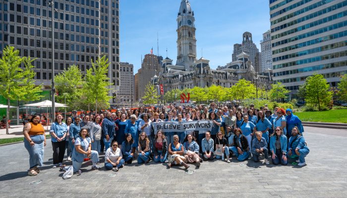 A smiling group wearing denim poses with a banner that says "Believe Survivors" in Love Park