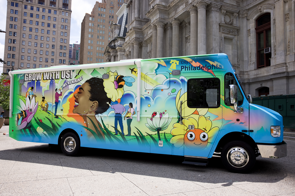 Image of the City's Hiring Bus parked in front of City Hall. The hiring bus is painted in a colorful mural with messages like "grow with us"