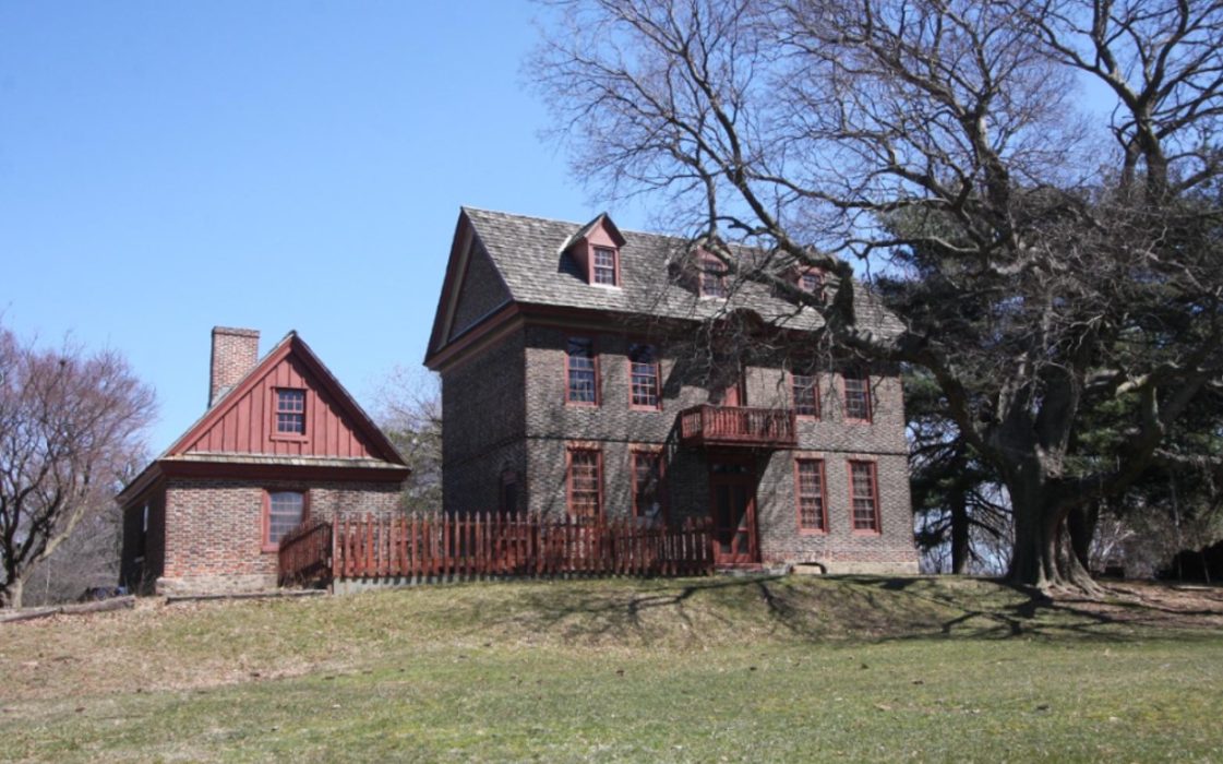 A photo of the Bellaire Manor in early spring, before the large oak tree in front of the house leafs out. The Georgian style home is made of brick. It has a small balcony on the second floor. An outbuilding has matching brickwork and red siding