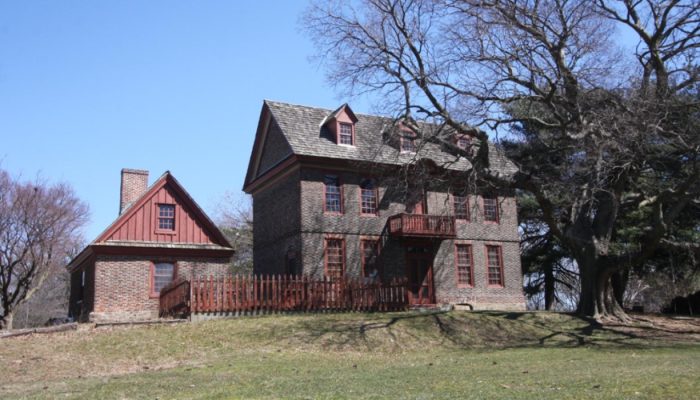 A photo of the Bellaire Manor in early spring, before the large oak tree in front of the house leafs out. The Georgian style home is made of brick. It has a small balcony on the second floor. An outbuilding has matching brickwork and red siding