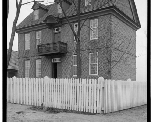 A black and white photo of a brick house with a white picket fence in the foreground.