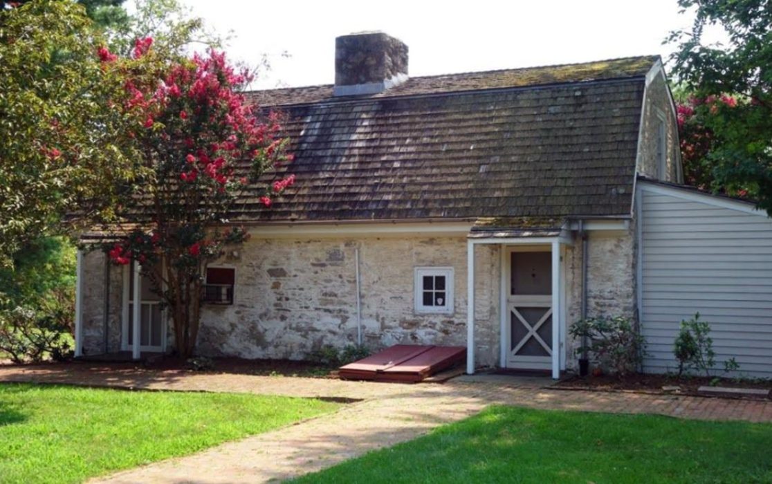 A small stone building with a gambrel roof. A flowering bush stands in front of it and brick footpaths cross in the foreground.