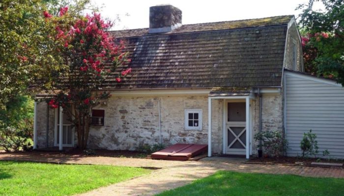 A small stone building with a gambrel roof. A flowering bush stands in front of it and brick footpaths cross in the foreground.