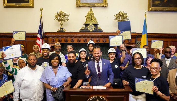 A group of men and women hold paper certificates above their head while gathered behind a podium with the City of Philadelphia seal on the front during a press conference in City Hall.