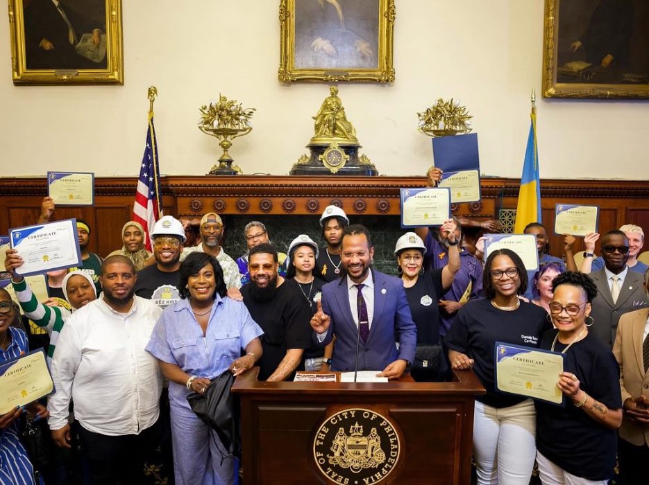 A group of men and women hold paper certificates above their head while gathered behind a podium with the City of Philadelphia seal on the front during a press conference in City Hall.