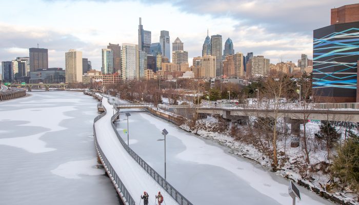 Two people cross country skiing on a snowed over Schuykill River Trail. The river is frozen over below the trail.