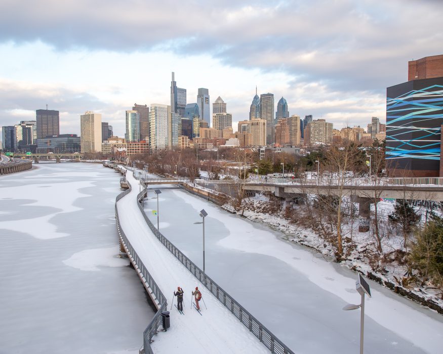 Two people cross country skiing on a snowed over Schuykill River Trail. The river is frozen over below the trail.