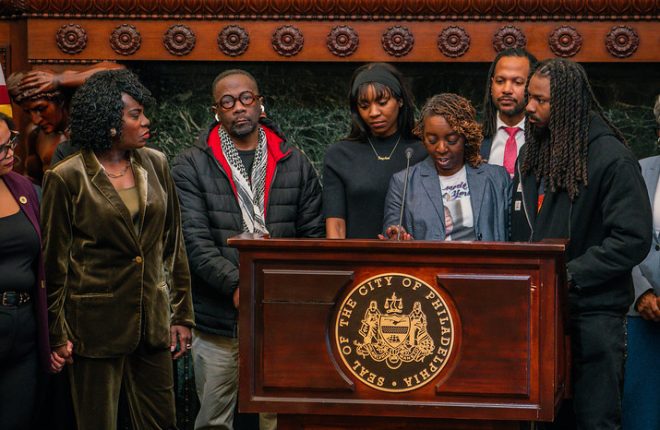 A group of men and women stand behind a podium with the City of Philadelphia seal on the front during a press conference at City Hall.