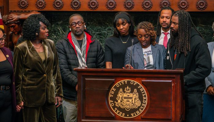A group of men and women stand behind a podium with the City of Philadelphia seal on the front during a press conference at City Hall.
