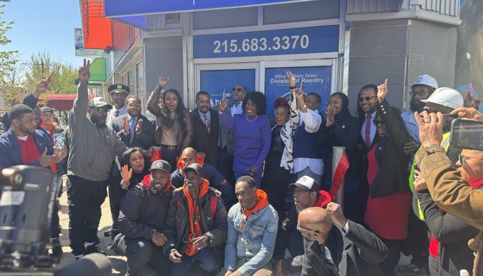 A group of people cut a red ribbon in front of a new facility with a blue sign, blue doors and grey walls.