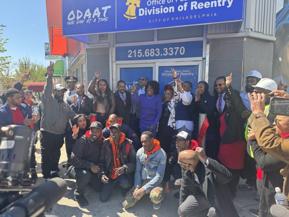 A group of people cut a red ribbon in front of a new facility with a blue sign, blue doors and grey walls.