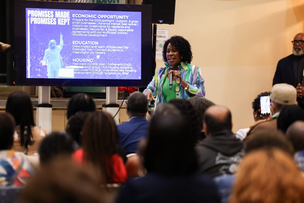 Mayor Cherelle L. Parker speaks into a microphone at a community budget town hall, addressing a seated audience with a presentation screen behind her highlighting economic opportunity, education, and housing priorities