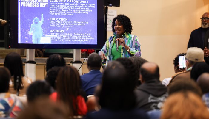 Mayor Cherelle L. Parker speaks into a microphone at a community budget town hall, addressing a seated audience with a presentation screen behind her highlighting economic opportunity, education, and housing priorities