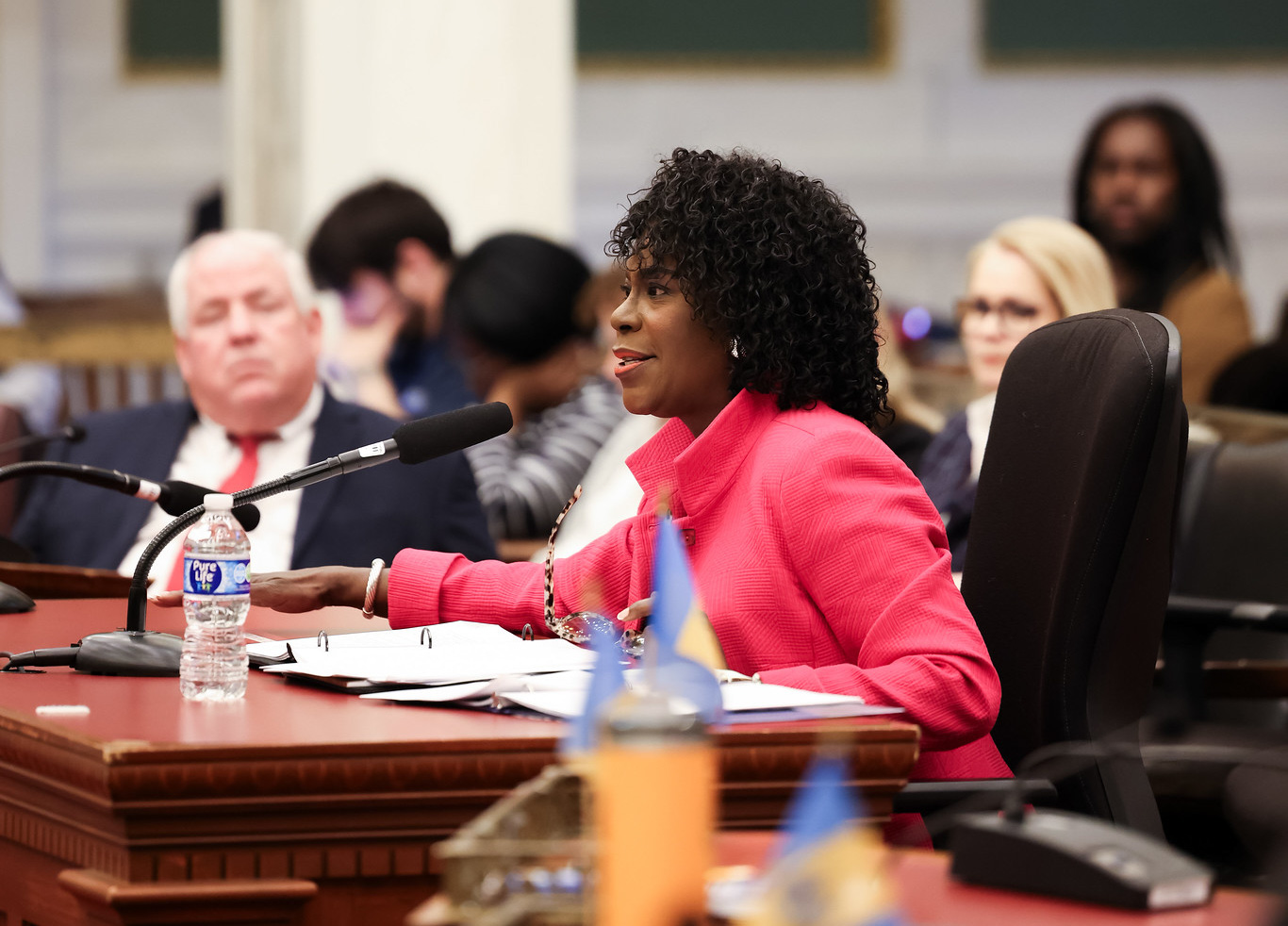 Mayor Cherelle L. Parker testifies before Philadelphia City Council, seated at a hearing table with a microphone, speaking while council members and attendees listen in the background.