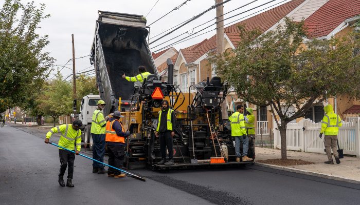 Uma equipe de pavimentação colocando asfalto em uma rua residencial como parte de um projeto de pavimentação. Os trabalhadores estão espalhando asfalto fresco usando vassouras quando ele sai de um caminhão basculante.