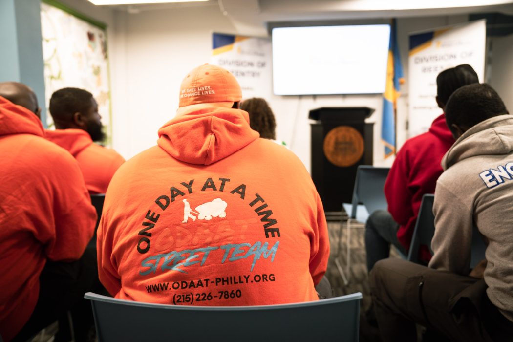 Individuals sit in front of a podium to listen to an event about justice-impacted people and voting. A man's sweatshirt says 'One Day at a Time'